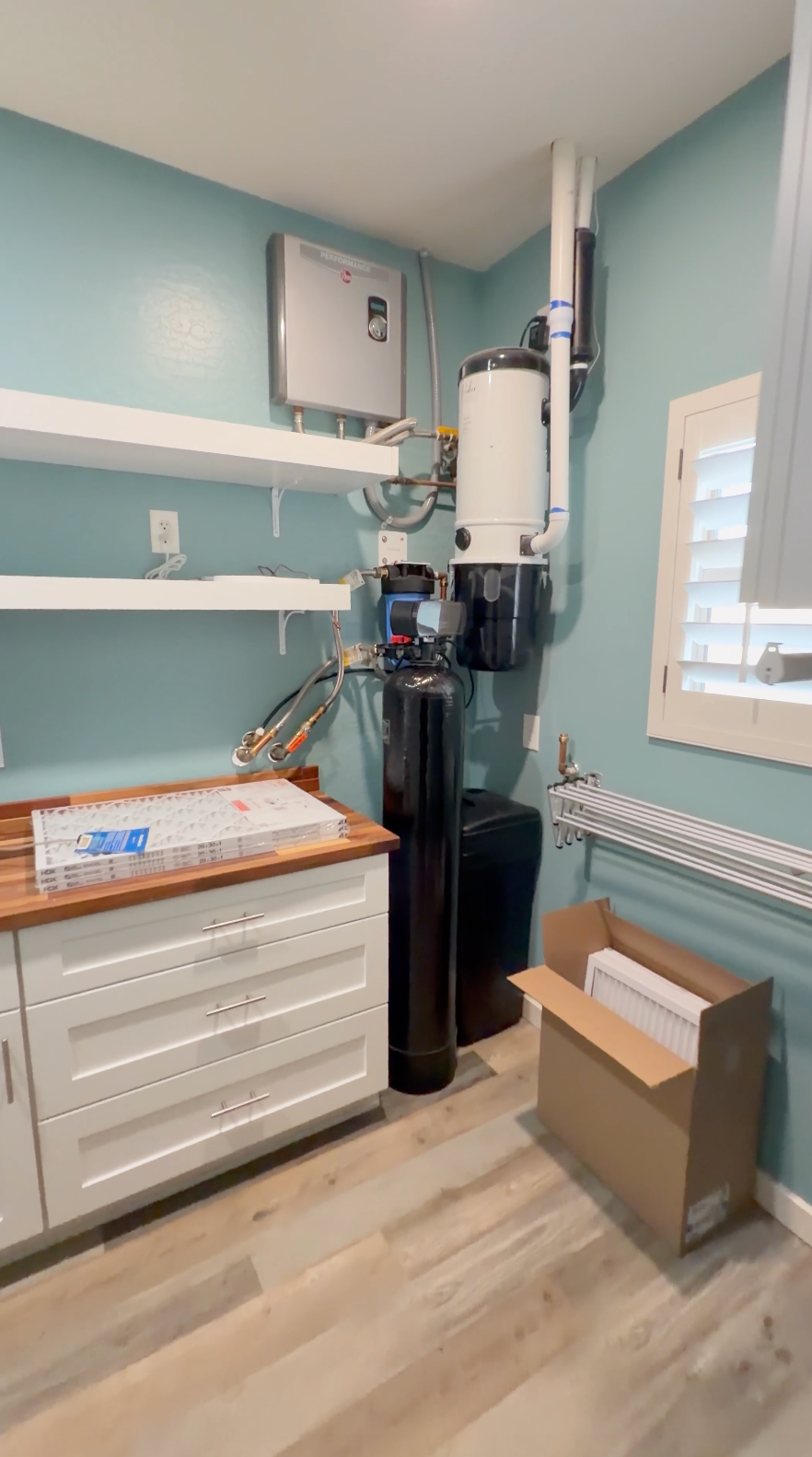 A residential water heater and tank system installed in a utility room with light blue walls, white cabinets, and wood-look flooring. The system includes a white water heater tank, a black cylindrical tank, metal piping, a gas furnace, and a cardboard box on the floor.