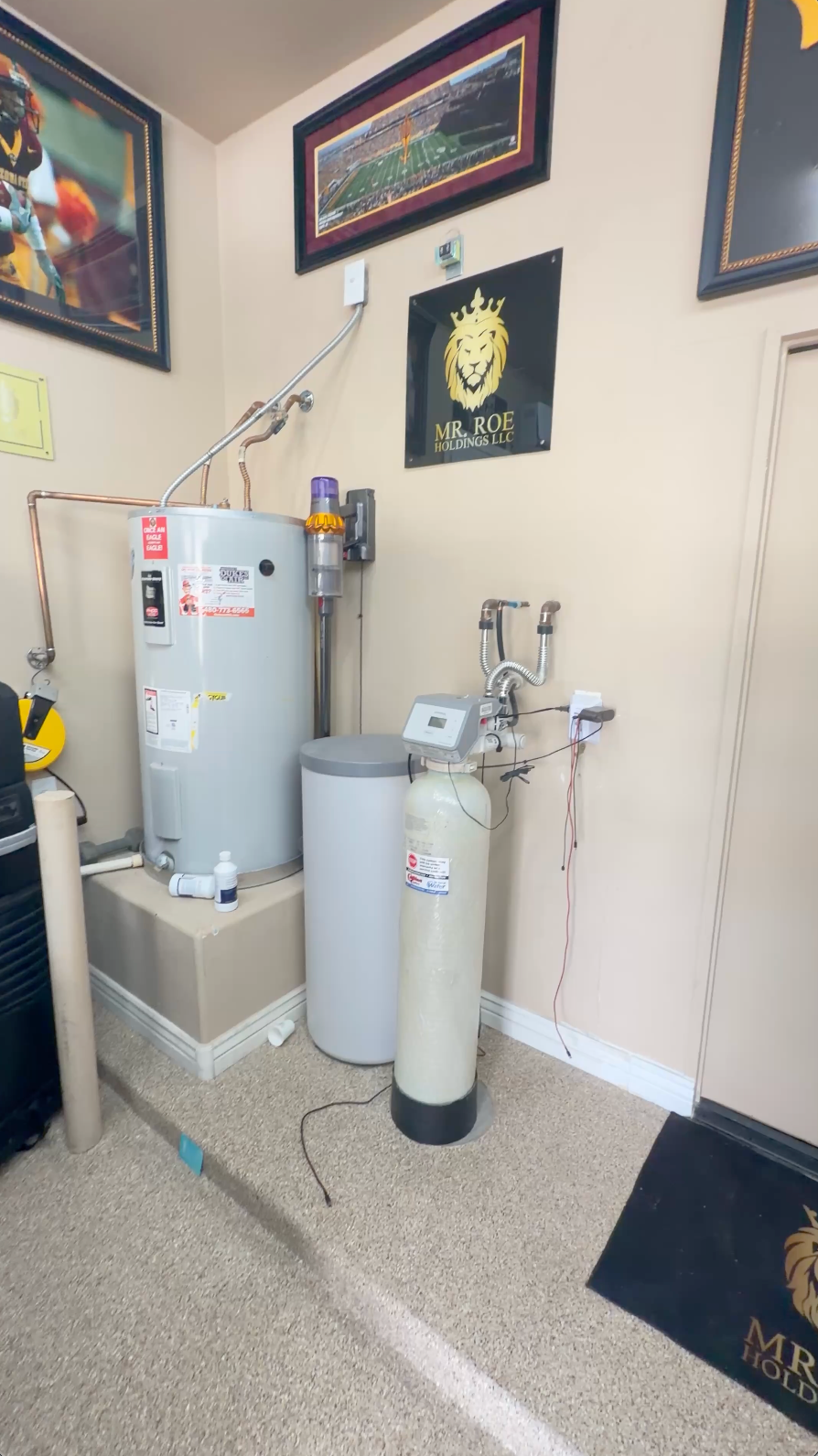 Water heater and water filter system installed in a home utility room with beige walls, framed pictures, and a black MR. ROE doormat visible in the corner.